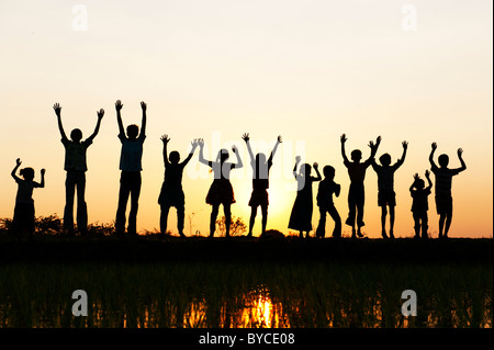 Silhouette Indian ragazze e ragazzi saltando e agitando in piedi su un riso risaie al tramonto. Andhra Pradesh, India Foto Stock