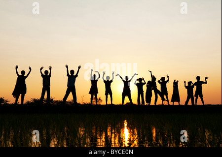 Silhouette Indian ragazze e ragazzi saltando e agitando in piedi su un riso risaie al tramonto. Andhra Pradesh, India Foto Stock