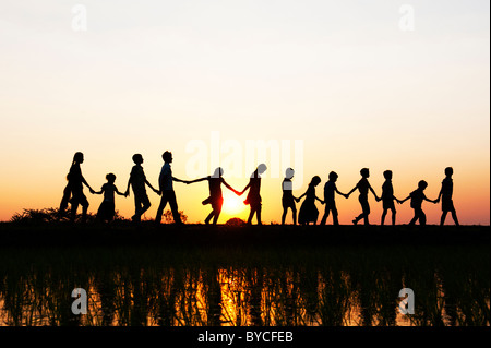 Silhouette Indian ragazze e ragazzi tenendo le mani a casa a piedi attraverso un riso risaie al tramonto. Andhra Pradesh, India Foto Stock