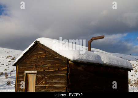 Vecchio e storico cabina con la neve sul tetto in inverno le montagne rocciose Montana USA Foto Stock