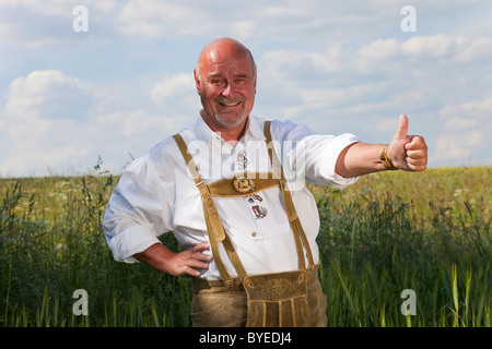 Uomo anziano indossando tradizionali bavaresi lederhosen, stando in piedi in un cornfield, pollice in alto, sorridente Foto Stock