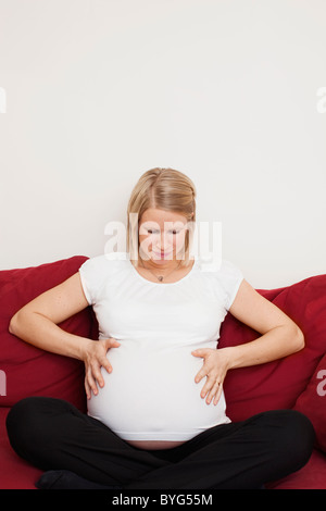 Donna incinta seduta sul divano con le mani sulla pancia Foto Stock