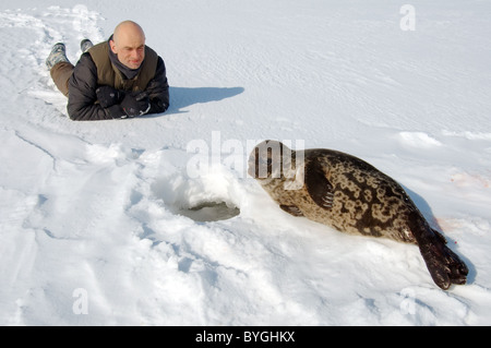 L uomo giace sulla neve e guarda alla guarnizione inanellato vicino a ice-foro. Guarnizione Jar, netsik o nattiq (Pusa hispida), Mare Bianco, artiche, Russia Foto Stock