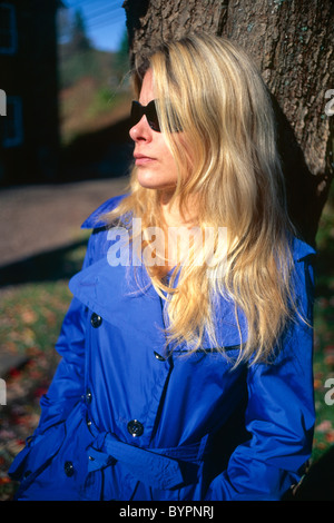 Giovane donna bionda in Blue Coat indossando occhiali da sole, appoggiata contro un albero, caduta, New Jersey Foto Stock