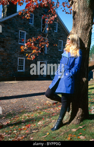 Giovane donna bionda in Blue Coat indossando occhiali da sole, appoggiata contro un albero, caduta, New Jersey Foto Stock