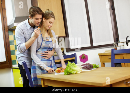 Caucasian donna bionda la preparazione di insalata in cucina, con l'uomo mangiare tomatoe slice e abbracciando il suo. Foto Stock