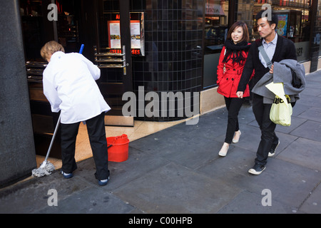 Giovani cinesi giovane a piedi passato una donna pavimento con straccio di Angus Steak House di Soho, su Charing Cross Road, Londra centrale. Foto Stock