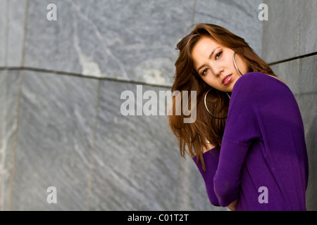 Giovane donna in posa di un vestito viola, appoggiata contro un muro grigio Foto Stock
