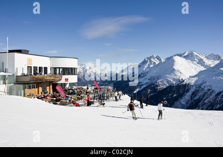 St Anton am Arleberg, Tirolo, Austria. Gli sciatori sulle piste da Galzig gondola stazione di vertice con la gente a prendere il sole fuori del ristorante Foto Stock