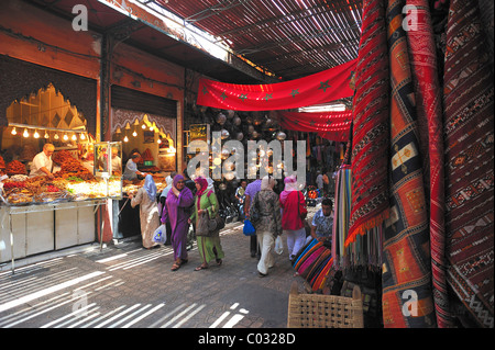 Strada nel mercato souk, bazaar nella Medina di Marrakech, un commerciante appendere fuori la sua mercanzia, opposta, un venditore di caramelle Foto Stock