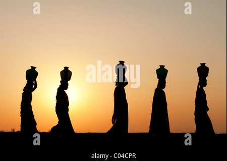 Il gruppo di donne indiane a piedi al tramonto portando vasi d'acqua sulle loro teste nella campagna indiana. Silhouette Foto Stock