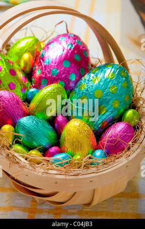 Tradizionale cestello in legno riempita con colorati stagnola avvolto le uova di Pasqua di cioccolata sul giallo e tovaglia bianca Foto Stock