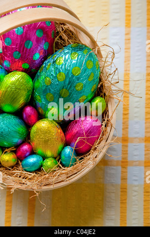 Tradizionale cestello in legno riempita con colorati stagnola avvolto le uova di Pasqua di cioccolata sul giallo e tovaglia bianca Foto Stock
