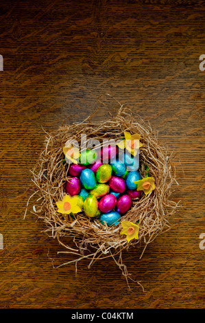 Gruppo di stagnola avvolto di piccole uova di pasqua di cioccolato in vari colori seduta nel fieno di un nido di uccelli su un tavolo in legno di quercia Foto Stock