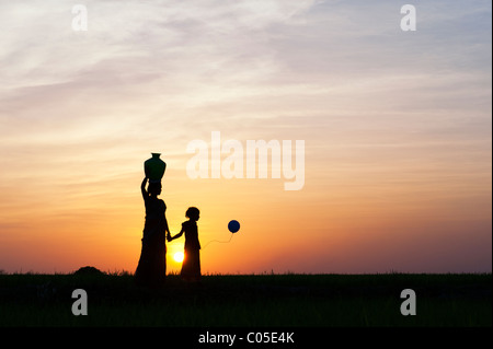 Madre indiana portando acqua pot con bambino e palloncini in campagna indiana. Silhouette Foto Stock