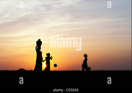 Madre indiana portando acqua pot con bambini e palloncini in campagna indiana. Silhouette Foto Stock