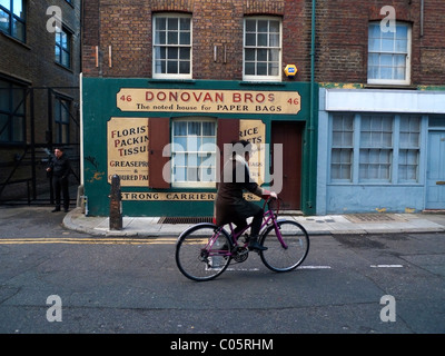 Una donna su una bicicletta Ciclismo passato Donovan Bros sacchetto di carta family business negozio Spitalfields in East London REGNO UNITO KATHY DEWITT Foto Stock