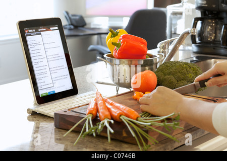 La donna la cucina in cucina mentre si seguono le istruzioni di cottura online su un iPad Foto Stock