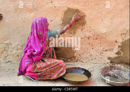 Anziani donna indiana intonacatura punti danneggiati del suo muro di casa con una miscela di sterco di vacca e la sabbia del deserto di Thar, Rajasthan Foto Stock