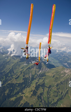 Skydivers di arancio e giallo tubo di aria sono caduta attraverso il cielo sopra le montagne verdi e le nuvole. Foto Stock