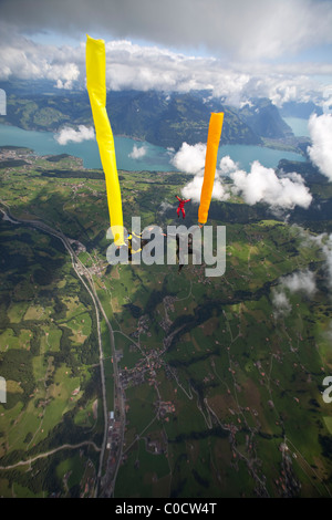 Skydivers di arancio e giallo dei tubi di aria vengono a cadere attraverso il cielo insieme su laghi e campi verdi. Foto Stock