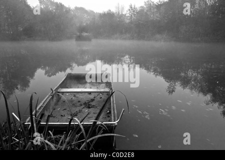 Autumn Fall paesaggio oltre il lago all'alba con nebbia appeso in aria e la vecchia barca a remi in primo piano Foto Stock