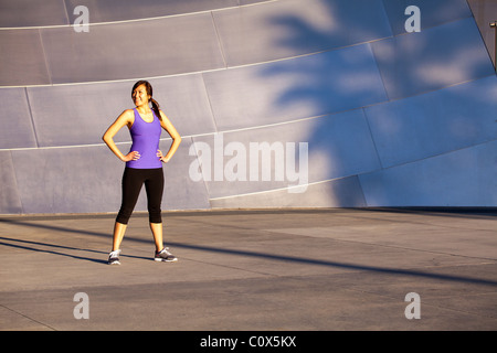 Americano asiatico femminile in appoggio in piedi di fronte a metal clad parete. Viola tank top e pantaloni neri. Foto Stock
