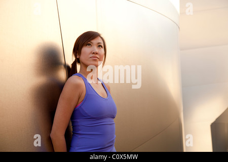 Americano asiatico femminile in appoggio appoggiata contro metal clad parete. Viola tank top e pantaloni neri. Foto Stock