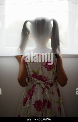 Bambina guarda fuori della finestra dietro tendina Foto Stock