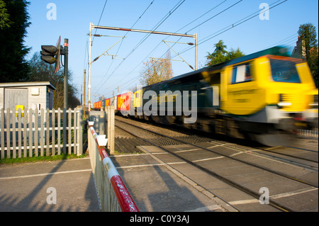 Lungo treno merci passa attraverso un livello senza equipaggio attraversando in Essex, UK. Le barriere sono in basso e la luce rossa di stop lampeggia. Foto Stock