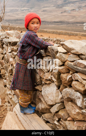 Giovane ragazzo bhutanesi sorge da un muro di pietra con un berretto in testa nelle zone rurali del Bhutan Foto Stock