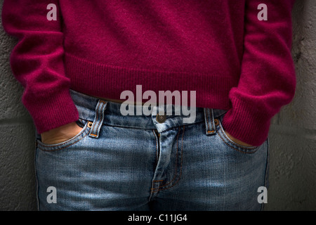 Una giovane donna indossa un rosso maglione di lana con jeans blu sorge con le mani nelle sue tasche. Foto Stock