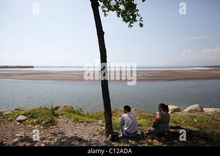 Un uomo e una donna sedere all'ombra di un albero che si affaccia sulla spiaggia e l'ingresso al porto in Quepos, Costa Rica Foto Stock