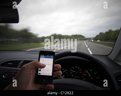 Un auto autista guida mentre utilizzando un dispositivo mobile / cellulare / smart phone. Driver utilizzando Facebook sul telefono. Foto Stock