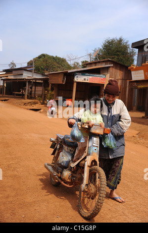 Donna spingendo un giovane bambino su una moto su una sabbiosa strada polverosa nel villaggio Bousra, zone di Mondulkiri, Cambogia Foto Stock
