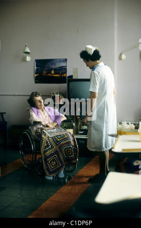 Una paziente anziana con una scialle accovacciata seduta su una sedia a rotelle che parla con un'infermiera in una stanza da giorno NHS all'ospedale Bronglais in 1980s 1980 Galles UK KATHY DEWITT Foto Stock