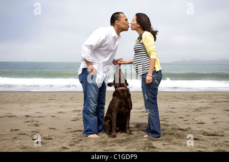 Il marito baciare moglie incinta sulla spiaggia mentre gli orologi del cane Foto Stock