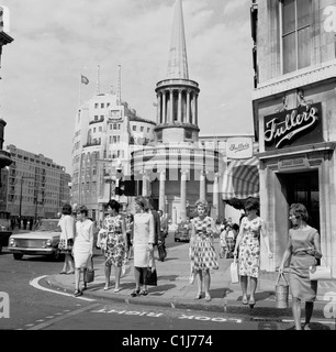 Anni '1960, donne in piedi su un marciapiede che stanno per attraversare una strada in Regent Street, Londra, All Souls Church e Broadcasting House, casa della BBC, dietro. Foto Stock
