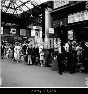 Anni '1950, assaggiatori ferroviari, con bagagli, coda per i biglietti nell'atrio della stazione ferroviaria di Victoria, sotto un cartello che promuove le case sul mare. Foto Stock