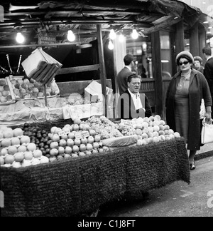 Anni '1960, storica, una tradizionale bancarella di frutta all'aperto, "J. Leonard"," a Leicester Square a Londra, con commercianti di mercato maschile e clienti femminili. Foto Stock