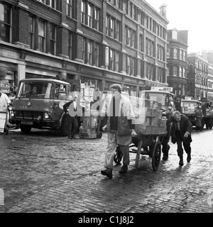 Anni '1950, un facchino che lavora al famoso mercato del pesce di Billingsgate a Londra, tira un carrello carico pesante su una strada acciottolata, con una spinta dal retro! Foto Stock