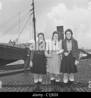 Anni '1950, tre giovani lavoratori portuali con gonne e sciarpe per la testa si posano insieme su una banchina acciottolata vicino al fiume Tyne, Newcastle-upon-Tyne, Inghilterra, Regno Unito. Foto Stock