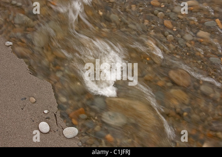 Questa natura foto mostra un primo piano della setosa acqua che scorre sulle rocce, con una spiaggia sabbiosa e frastagliate ledge tenendo un paio di ciottoli. Foto Stock