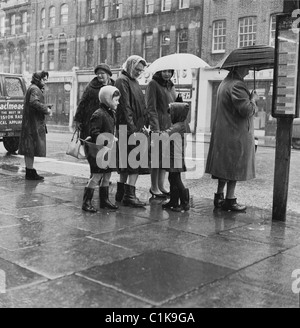 1960s, ad una fermata dell'autobus, una madre con i suoi due figli piccoli in attesa con altre Signore su un marciapiede sotto la pioggia per un autobus, Londra, Inghilterra, Regno Unito. Foto Stock