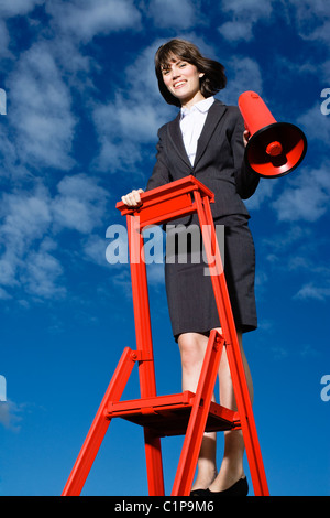 Sorridente imprenditrice permanente sulla parte superiore della scaletta rosso contro il cielo blu e rosso di contenimento megafono Foto Stock