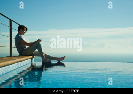 Uomo seduto sul bordo della piscina a sfioro, la messaggistica di testo con un telefono cellulare Foto Stock