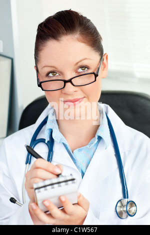 Riflessivo medico donna sorridente verso la telecamera tenendo un blocco note nel suo ufficio seduto Foto Stock