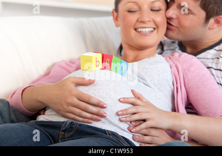 Giovane uomo baciare sua moglie incinta con il bambino lettere sul ventre sdraiato sul divano Foto Stock