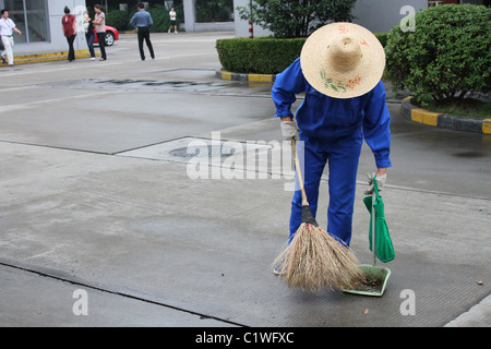 Donna che indossa cappello di paglia spazzando un parcheggio con una scopa di paglia. Shanghai, Cina. Foto Stock