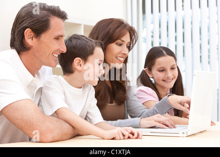 Un attraente felice, sorridente famiglia della madre, padre e figlio e figlia seduti ad un tavolo utilizzando un bianco computer portatile Foto Stock